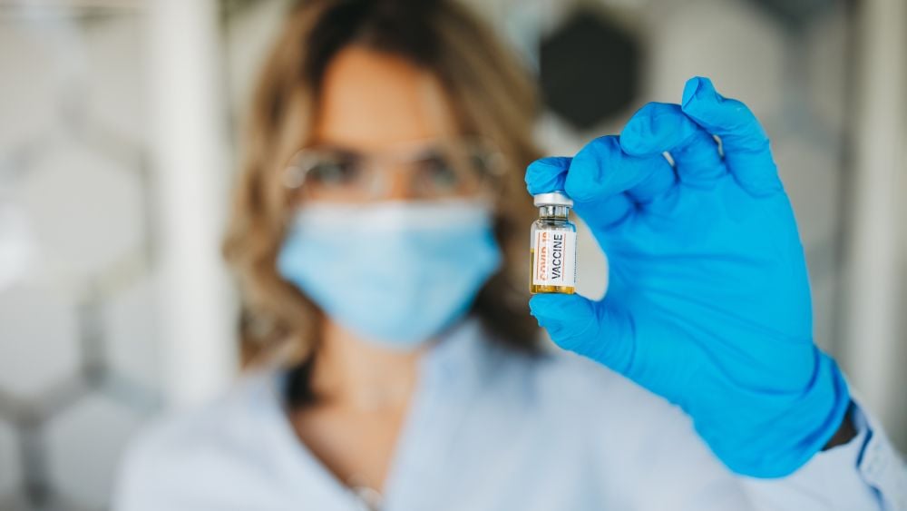 Person wearing a face mask and gloves holding up a vaccine vial to the camera